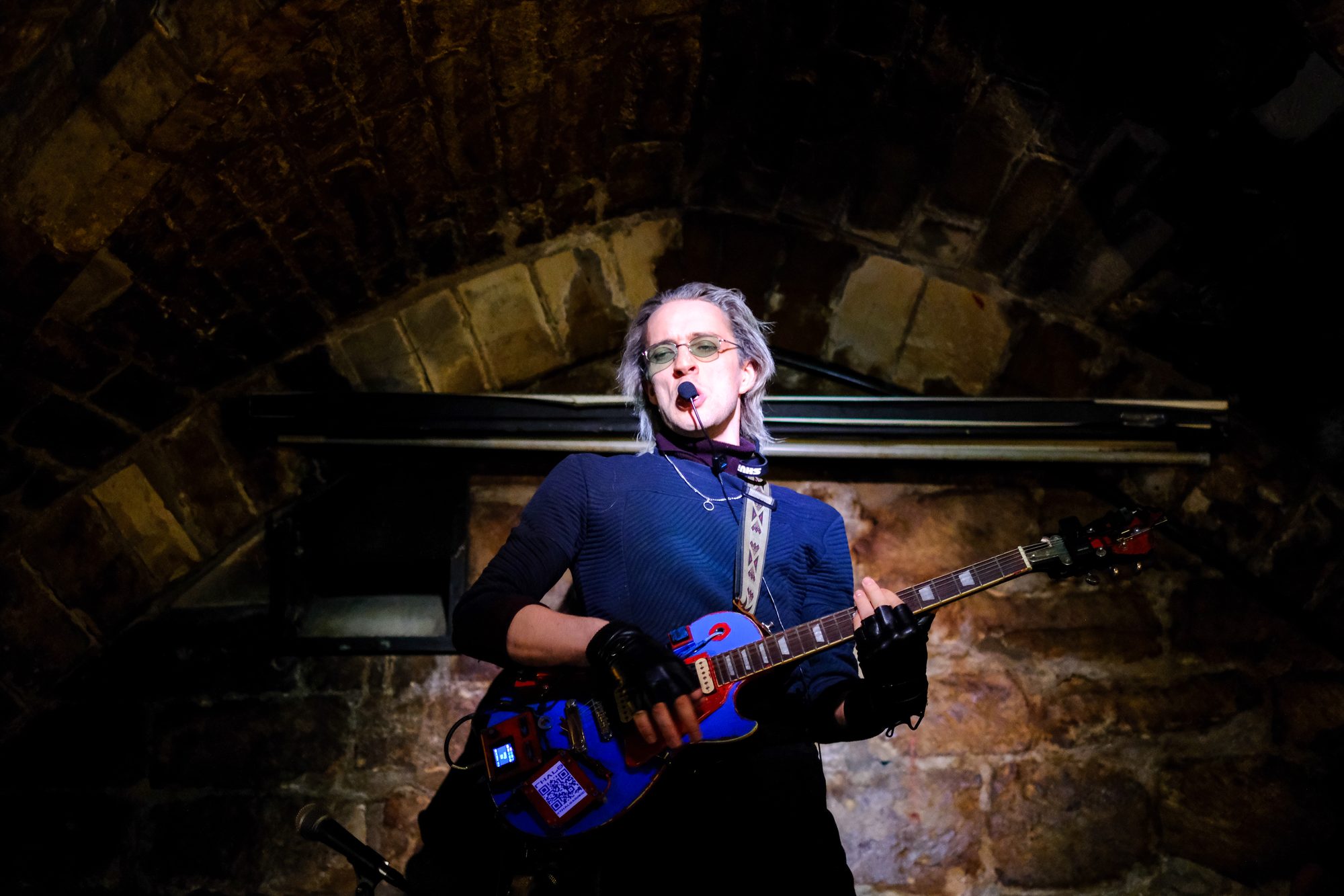 Thalo on stage in a stone cellar in Paris, blue jumper, red and blue guitar